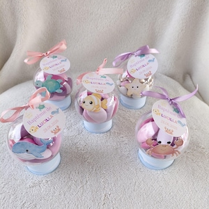 Peut inclure: Des contenants sphériques transparents remplis de bonbons roses et de décorations de créatures marines. Chaque contenant a une base bleu clair, un ruban et une étiquette "Baptême de LUCILLA 28.06.2026".