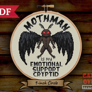 May include: A cross-stitch hoop featuring a pixelated Mothman design with red eyes and black wings. Text reads "Mothman is my Emotional Support Cryptid." The hoop is 9 inches in diameter and has a wooden frame. The PDF logo is in the upper left corner.