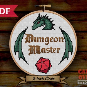 Puede incluir: Un aro de bordado blanco con un dragón verde, un dado rojo de 20 caras y el texto "Dungeon Master" en dorado. El aro tiene un diámetro de 20 cm.