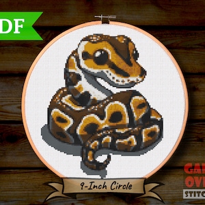 May include: A cross-stitch pattern of a curled up, brown and white snake with large eyes. The pattern is in a 9-inch embroidery hoop and is labelled "Game Over Stitches".