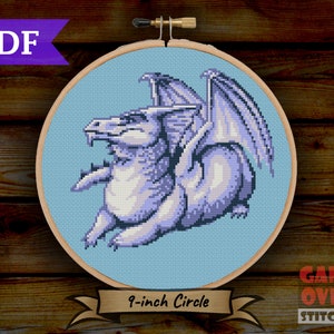 May include: A cross-stitch pattern of a light blue dragon with wings folded behind it. The pattern is in a 9-inch embroidery hoop. The text "GAME OVER STITCHES" is in the lower right corner of the image.