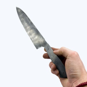 K&ouml;nnte beinhalten: Eine Hand h&auml;lt ein langes, silbernes Messer mit einer strukturierten Klinge und einem schwarzen Griff. Das Messer ist unvollendet und hat ein raues, industrielles Aussehen.