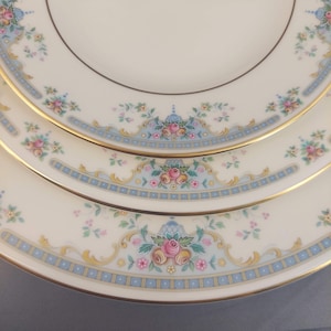 May include: Close-up of three white porcelain plates with a floral design. The plates have a light blue border with a repeating square pattern and gold trim. Pink and yellow roses are the focal point of the design.
