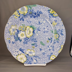 May include: A round decorative plate with a scalloped edge. The plate has a blue and white floral pattern with yellow and white flowers. The plate is supported by a white stand.