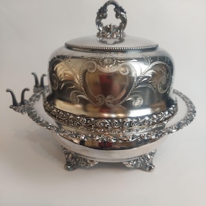 May include: A silver butter dish with a lid and intricate floral patterns. The dish features a round base with decorative feet and a matching lid with a handle. The rim of the dish has a detailed, scalloped edge.