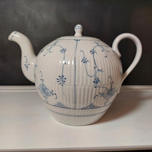 Może przedstawiać: Biały porcelanowy imbryk z niebieskim kwiatowym wzorem. Imbryk ma okrągły korpus z pionowymi żebrowanymi detalami, zakrzywioną wylewkę i uchwyt w kształcie pętli. Pokrywka zwieńczona jest małym guzikiem. Niebieski kwiatowy wzór zawiera kwiaty i winorośl.