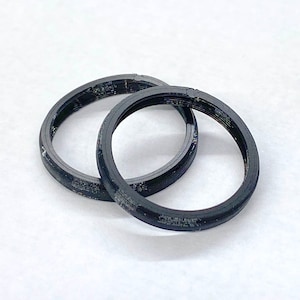 Puede incluir: Dos objetos circulares negros con un diseño en capas se colocan sobre una superficie blanca. Los objetos parecen ser anillos o bandas, posiblemente hechos de un material compuesto. La iluminación es brillante y uniforme, resaltando la textura.