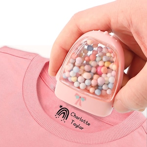 May include: Pink fabric stamp with a clear container filled with colorful beads. The stamp is being used to stamp a pink shirt with a rainbow, crown, and the text "Charlotte Taylor".