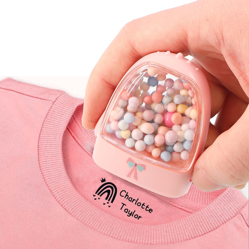 May include: Pink fabric stamp with a clear container filled with colorful beads. The stamp is being used to stamp a pink shirt with a rainbow, crown, and the text "Charlotte Taylor".
