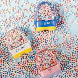 May include: Three colorful toy containers filled with pastel-colored foam balls. The containers are shaped like small vehicles and have different messages on the front: "be yourself", "be brave", and a pink container with a bow.