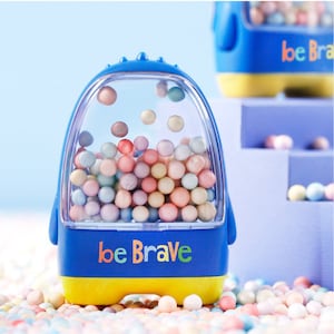 May include: A blue and yellow plastic toy with a clear plastic container filled with colorful balls. The toy has the words "be Brave" printed on the front.