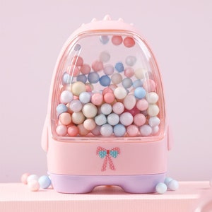 May include: A pink plastic toy shaped like a gumball machine with a clear plastic window filled with colorful pastel beads. The toy has a purple base and a pink bow on the front.