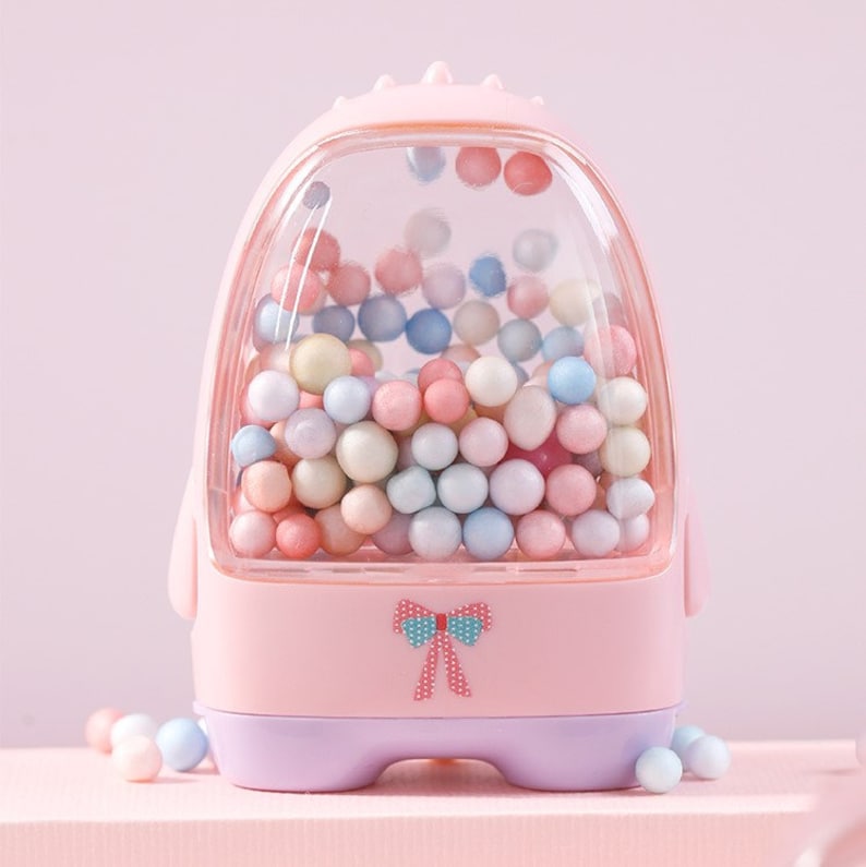 May include: A pink plastic toy shaped like a gumball machine with a clear plastic window filled with colorful pastel beads. The toy has a purple base and a pink bow on the front.