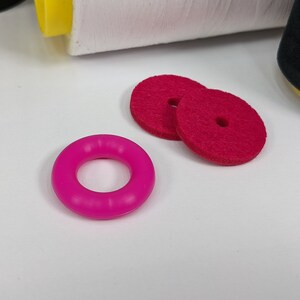 以下が含まれることがあります： 中央に穴が開いたピンクのフェルトの円形と、ピンクのゴム製のリング。円形は白い表面の上に置かれています