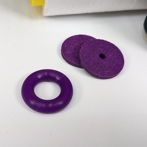 Puede incluir: Tres canillas de máquina de coser de color morado, dos son planas y una tiene forma de anillo.