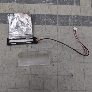 May include: A small, clear plastic light fixture with a silver foil backing and a black wire with a white connector. The light fixture is on a gray surface with a ruler.