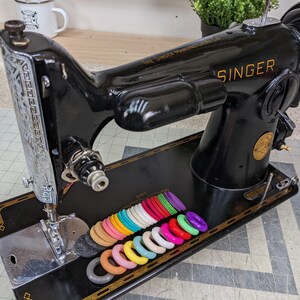 Op de afbeelding: Een zwarte Singer naaimachine met een zilveren voetpedaal en een set kleurrijke plastic ringen op de tafel. De machine heeft een gouden naamplaatje met de tekst "The Singer Manufacturing Co. U.S.A." en "SINGER".