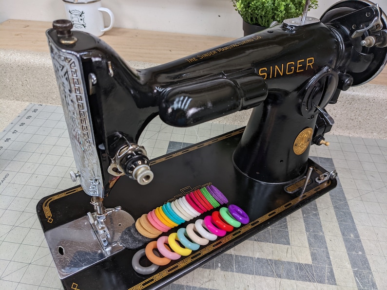 Op de afbeelding: Een zwarte Singer naaimachine met een zilveren voetpedaal en een set kleurrijke plastic ringen op de tafel. De machine heeft een gouden naamplaatje met de tekst "The Singer Manufacturing Co. U.S.A." en "SINGER".