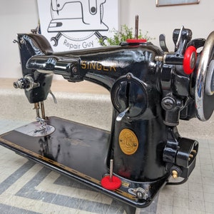 Op de afbeelding: Een zwarte Singer naaimachine met een gouden naamplaatje en rode accenten. De machine heeft een voetpedaal en een naald.