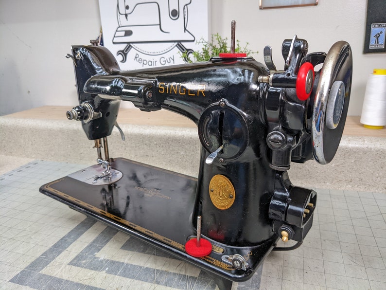Op de afbeelding: Een zwarte Singer naaimachine met een gouden naamplaatje en rode accenten. De machine heeft een voetpedaal en een naald.