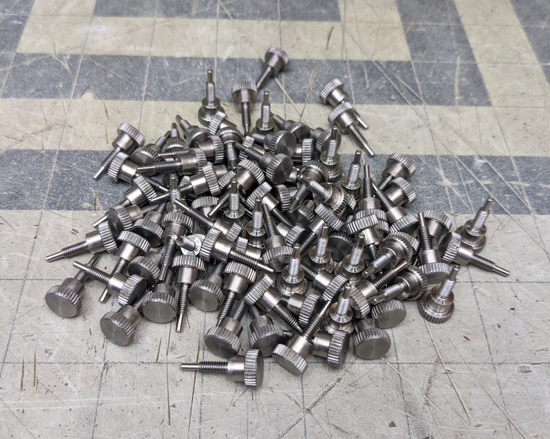 May include: A large pile of silver-colored metal screws with knurled heads. The screws are arranged in a random pattern and are all the same size.