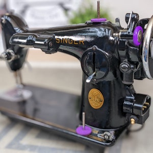 Op de afbeelding: Een zwarte Singer naaimachine met een gouden naamplaatje en een gouden rond embleem. De machine heeft een voetpedaal en een stroomkabel.
