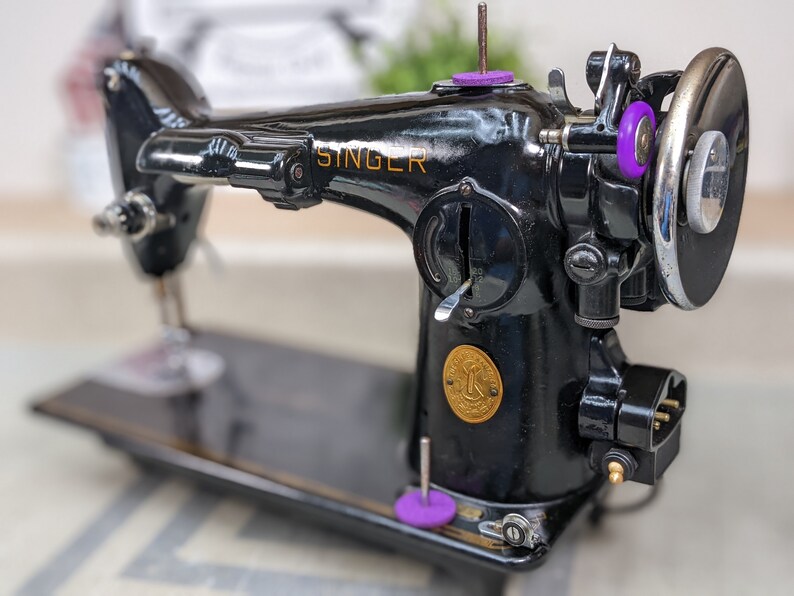 Op de afbeelding: Een zwarte Singer naaimachine met een gouden naamplaatje en een gouden rond embleem. De machine heeft een voetpedaal en een stroomkabel.