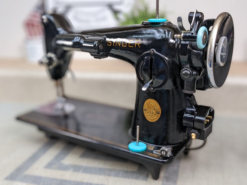 Op de afbeelding: Een zwarte Singer naaimachine met een gouden logo en een voetpedaal. De machine heeft een naald en draad op zijn plaats, en de spoel is zichtbaar.