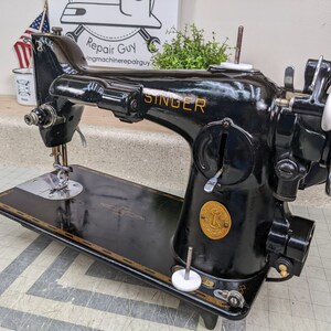 Op de afbeelding: Een zwarte Singer naaimachine met een gouden naamplaatje en een wit voetpedaal. De machine heeft een groot handwiel en een zichtbare naald. De machine staat op een zwart oppervlak.