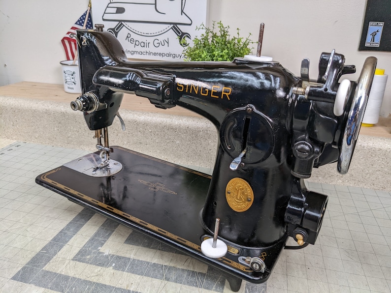 Op de afbeelding: Een zwarte Singer naaimachine met een gouden naamplaatje en een wit voetpedaal. De machine heeft een groot handwiel en een zichtbare naald. De machine staat op een zwart oppervlak.