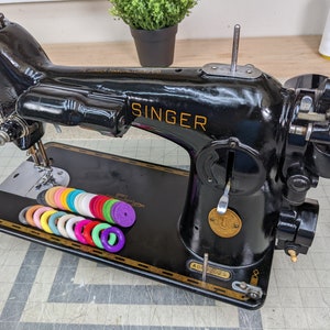 Op de afbeelding: Een zwarte Singer naaimachine met gouden accenten en een witte spoelhouder. De machine heeft een naald en een voetpedaal. Er liggen kleurrijke spoelen op de tafel voor de machine.