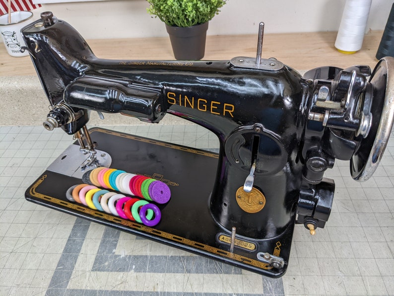 Op de afbeelding: Een zwarte Singer naaimachine met gouden accenten en een witte spoelhouder. De machine heeft een naald en een voetpedaal. Er liggen kleurrijke spoelen op de tafel voor de machine.