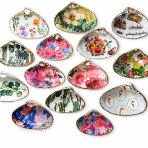 May include: A collection of thirteen decorative clam shells, each featuring unique decoupage designs including floral patterns, cacti, botanical greenery, and architectural illustrations, all finished with a gold-painted edge.