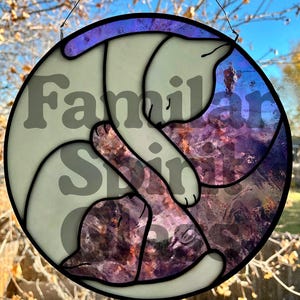 May include: A round stained glass piece featuring two stylized cats in a yin-yang pose. The cats are white with black outlines, and the background is a mix of purple, blue, and brown. The words "Familiar Spirit" are overlaid.