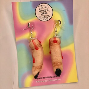Pode incluir: Um par de brincos em forma de dedos ensanguentados com esmalte preto. Os brincos estão em um cartão com fundo arco-íris e o texto "Wonderful Little Weirdos" e "Handmade in Manchester".