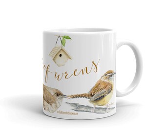 Puede incluir: Taza de cerámica blanca con una casita de pájaros marrón, dos pájaros marrones y el texto "A Song of Wrens" en escritura dorada.