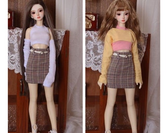 Puede incluir: Se muestran dos muñecas de moda, cada una con un atuendo diferente. Una muñeca tiene cabello castaño largo y un top corto blanco con una falda a cuadros. La otra muñeca tiene cabello rubio y un top corto amarillo con una camisa rosa y una falda a cuadros.