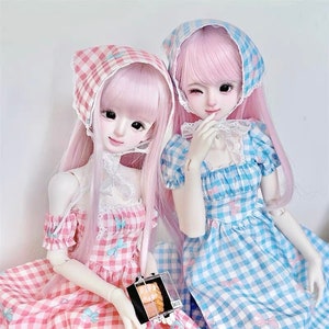 Könnte beinhalten: Zwei Puppen in rosa und blauen Gingham-Kleidern mit passenden Stirnbändern. Die Puppen haben rosa Haare und halten eine kleine Schachtel.