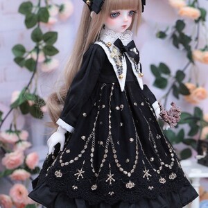 May include: A doll wearing a black dress with gold embroidery and a matching black headband. The dress features a white collar and cuffs, and a black bow. The doll has long blonde hair and is holding a small bouquet of flowers.