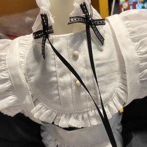May include: White dress with black ribbon bows and white lace trim. The dress has a white collar with a ruffled edge and two white buttons.