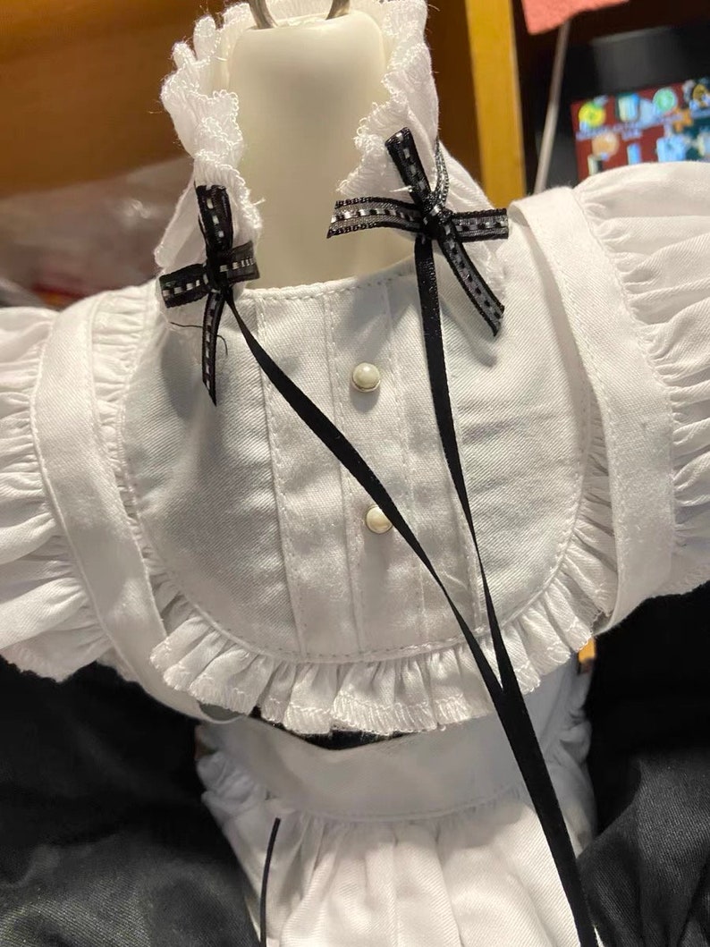 May include: White dress with black ribbon bows and white lace trim. The dress has a white collar with a ruffled edge and two white buttons.