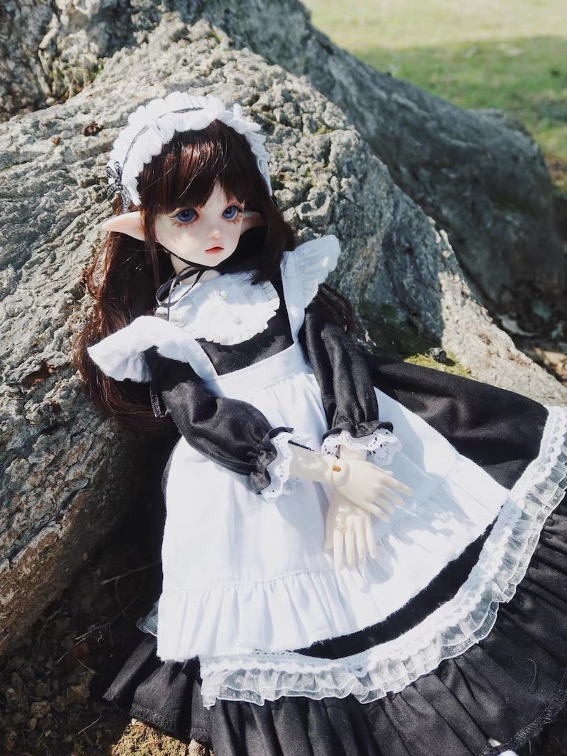 May include: A doll wearing a black and white maid outfit with a white lace collar and a white lace headband. The doll has long brown hair, blue eyes, and pointed ears. The doll is sitting on a large rock.