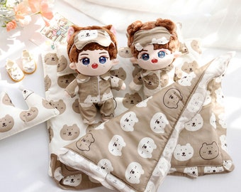 May include: Two plush dolls wearing brown pajamas and sleep masks are lying on a brown and white patterned blanket with a brown pillow. The blanket has a white border with a scalloped edge.