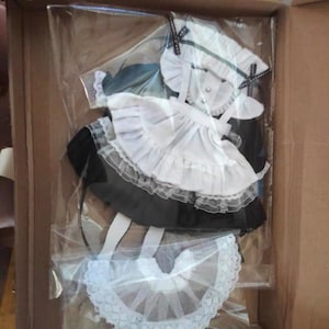May include: A white and black maid outfit for a doll. The outfit includes a white apron with black lace trim, a black skirt with white lace trim, a white collar with black bows, and a white headband with black bows.