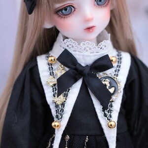 May include: A close-up of a doll with blue eyes and long, light brown hair. The doll wears a black dress with a white collar and a black bow. The dress is accented with gold-colored beads and a decorative pendant.