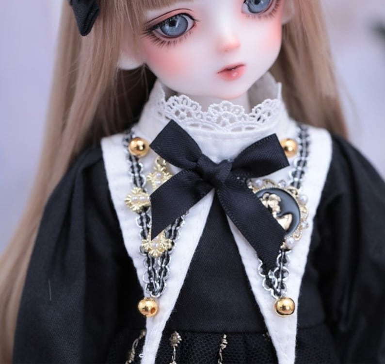 May include: A close-up of a doll with blue eyes and long, light brown hair. The doll wears a black dress with a white collar and a black bow. The dress is accented with gold-colored beads and a decorative pendant.