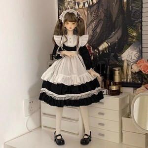 May include: A doll wearing a black and white maid outfit with a white apron and lace trim. The doll has brown hair and is wearing black shoes.