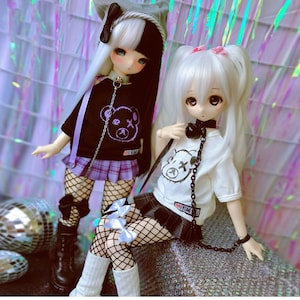 May include: Two dolls with white hair wearing black and white outfits with a bear graphic. The dolls are wearing fishnet stockings and have black chokers with chains.