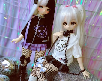 May include: Two dolls with white hair wearing black and white outfits with a bear graphic. The dolls are wearing fishnet stockings and have black chokers with chains.