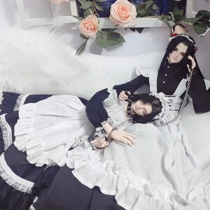 May include: Two dolls wearing maid outfits with black and white ruffles. One doll is wearing a black choker with a silver chain. The dolls are lying on a white bed with a white fluffy blanket.